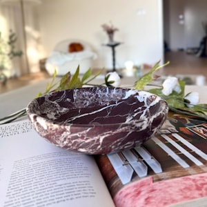 May include: A maroon marble bowl with white veining sits on an open book. The bowl is round and has a smooth, polished surface. White flowers and greenery are arranged around the bowl, adding a natural touch to the scene.