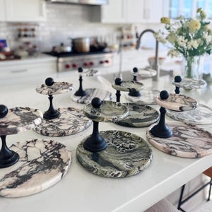 May include: A collection of tiered serving stands featuring circular marble bases and platforms in various shades of white, grey, and green with black veining. Each stand has a central black metal pedestal topped with a small black sphere.