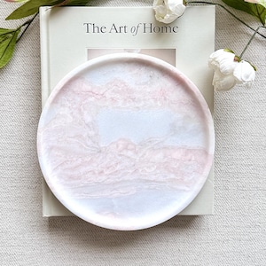 May include: A circular tray featuring a marbled pattern in soft shades of pink and white. The tray is positioned on top of a book titled The Art of Home, which is decorated with white floral stems and green leaves against a neutral background.