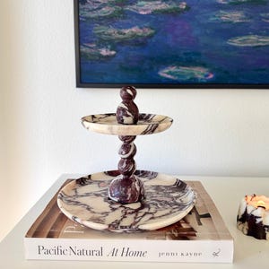 May include: A two-tiered serving stand crafted from white and deep purple marble. The stand features a central spiraled pillar connecting a smaller top tray and a larger bottom tray, both displaying a natural, swirling veined pattern throughout.