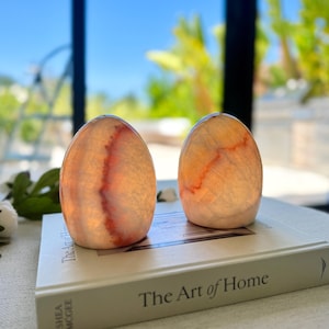 May include: Two smooth, egg-shaped stone sculptures with translucent, pale orange and white banding and deep red veining, resting on a white hardcover book titled The Art of Home by Shea McGee, featuring a flat base and rounded top.