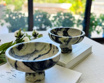 May include: Two small, circular bowls crafted from polished stone featuring a marbled pattern of cream, light green, and deep black veins. Each bowl is supported by a short, thick pedestal base, creating a raised, footed silhouette.