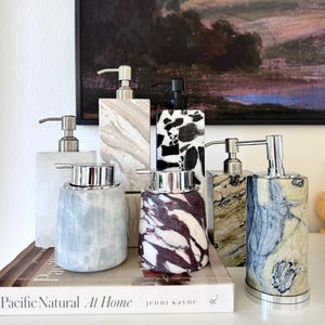 May include: A collection of six soap dispensers with pump mechanisms, featuring various stone textures and colours including white, beige, black and white terrazzo, deep purple, green, and blue-grey marble, arranged on a book titled Pacific Natural At Home.