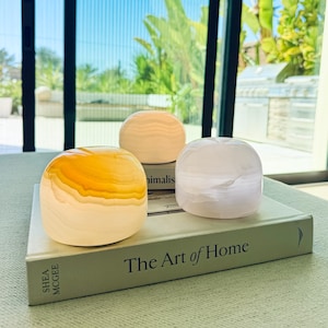 Puede incluir: Tres objetos de piedra lisos, redondeados y translúcidos con patrones de bandas en tonos amarillo, blanco y rosa pálido, sobre un libro de tapa dura titulado The Art of Home de Shea McGee, con una cubierta gris claro y texto en negro.