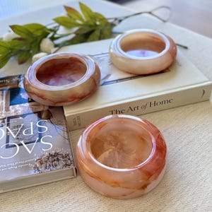 May include: Three decorative, round, resin bowls with a marbled effect in shades of peach, orange, and white. The bowls are arranged on a neutral surface with two books, one titled "The Art of Home".