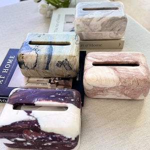 Può includere: Quattro scatole rettangolari in pietra per fazzoletti con angoli arrotondati e una fessura superiore. Ogni scatola presenta un motivo marmorizzato unico in diverse colorazioni: bianco e grigio, verde e blu, rosa e bianco, viola scuro e bianco.