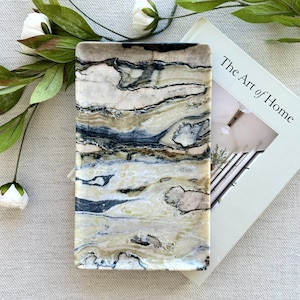 Puede incluir: Una bandeja rectangular de piedra con una superficie pulida con patrones de remolinos en crema, beige y gris carbón oscuro. La bandeja está colocada junto a un libro titulado The Art of Home y una pequeña etiqueta blanca con el número 0020.