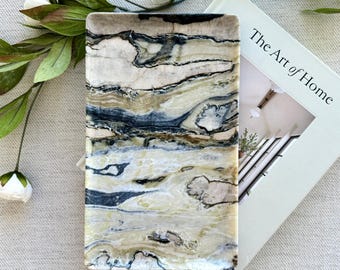 May include: A rectangular stone tray featuring a polished surface with swirling patterns of cream, beige, and dark charcoal gray. The tray is positioned next to a book titled The Art of Home and a small white tag with the number 0020 printed on it.