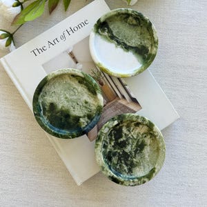 Puede incluir: Tres posavasos redondos de mármol verde y blanco con un patrón veteado natural. Los posavasos están colocados sobre un libro titulado "The Art of Home", con flores blancas y hojas verdes en el fondo.