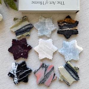 Peut inclure: Neuf dessous de verre en pierre en forme d'étoile disposés en grille, présentant divers motifs et couleurs naturels, notamment noir, blanc, rose, vert et rouge foncé. Les dessous de verre sont placés sous un livre blanc intitulé The Art of Home.