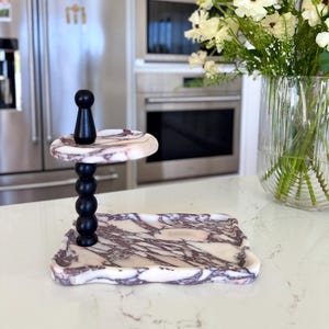 May include: A two-tiered serving stand featuring a rectangular base and a smaller circular top tray, both crafted from white marble with prominent purple veining. A central black wooden spindle with a spherical design connects the two tiers.