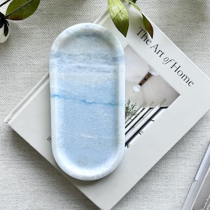 May include: A marble-like, oval tray with a blue and white color scheme. The tray is resting on a book titled "The Art of Home". Decorative greenery is in the background.