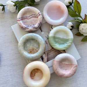 Puede incluir: Seis pequeños platos redondos de piedra en tonos rosa, verde y crema. Los platos tienen un acabado liso y pulido y diferentes patrones. Algunos tienen forma de cuenco, otros de rosquilla. Flores blancas y vegetación en el fondo.