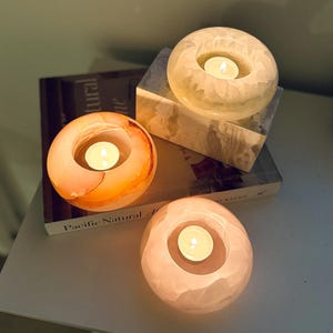 Puede incluir: Tres portavelas redondos de piedra pulida en tonos crema, naranja y rosa pálido, cada uno con una vela de té encendida. Los portavelas están dispuestos sobre un libro titulado Pacific Natural, que presenta un bloque de piedra veteada.