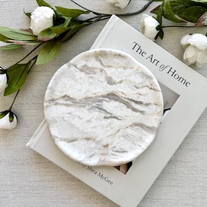 May include: A circular white marble tray with grey and beige veining patterns. The tray is resting on a white hardcover book titled The Art of Home by Shea McGee, accompanied by a decorative vine with white peony flowers and green leaves.