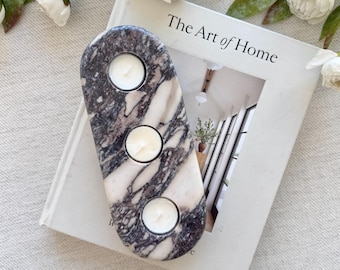 Puede incluir: Un portavelas de mármol con tres aberturas circulares, cada una con una vela blanca encendida. El soporte tiene un patrón veteado gris y blanco. Descansa sobre un libro titulado "The Art of Home" con flores blancas y vegetación en el fondo.