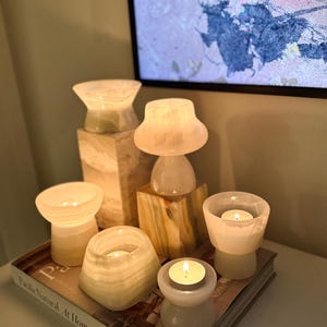May include: A collection of six cream-coloured stone candle holders with smooth, translucent surfaces, each containing a lit tea light candle. The holders are arranged on a book titled Pacific Natural At Home by Jenni Kayne, with two stone blocks.