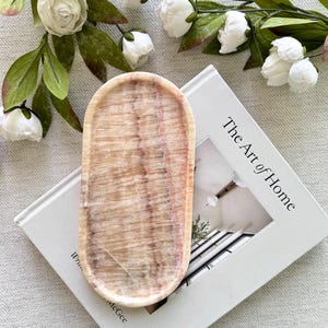 以下が含まれることがあります： ベージュと茶色の楕円形の大理石のトレイが、「The Art of Home」というタイトルの本の上に置かれています。 トレイは、研磨された仕上げの天然石素材で作られています。 白い花と緑の葉が背景にあります。