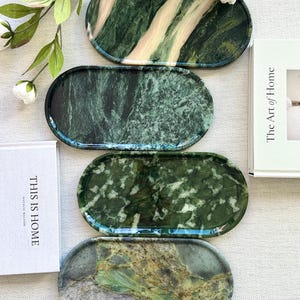 Pode incluir: Quatro tabuleiros de forma oval com superfícies lisas e polidas, apresentando padrões distintos de pedra natural em tons de verde floresta profundo, azeitona e creme. Cada tabuleiro exibe veios e texturas minerais únicos.