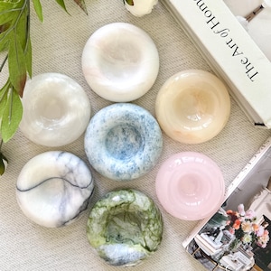 May include: A collection of seven circular stone bowls with smooth, polished surfaces. The bowls feature various colors including white, pale blue, soft pink, cream, and green, with some displaying natural veining and marbled patterns.