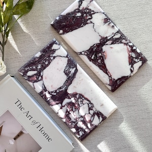 May include: Two rectangular marble slabs featuring a white base with deep purple and black veining patterns. Next to the slabs is a book with a white cover that has the text The Art of Home printed in black on the front cover.