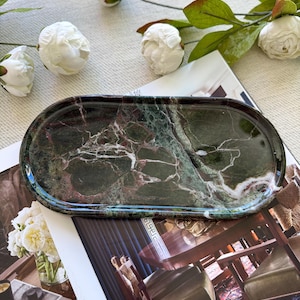 May include: An oval-shaped tray crafted from polished dark green marble featuring prominent white and deep red veining throughout the stone surface. The tray has a smooth, glossy finish and a raised rim, showcasing the natural patterns of the rock.