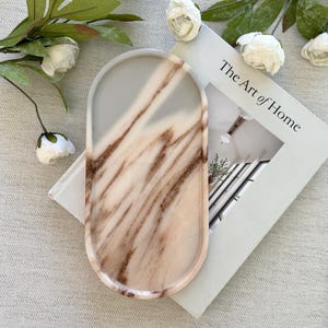 May include: An oval-shaped tray featuring a smooth, polished surface with a marbled pattern in shades of cream, beige, and brown. The tray has a raised edge and is positioned on top of a book titled The Art of Home, surrounded by white flowers.