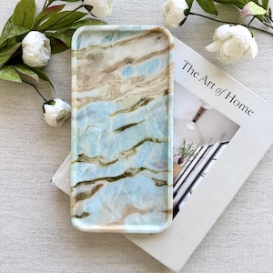 May include: A rectangular tray featuring a light blue and tan marbled pattern with smooth, rounded edges. The tray is positioned on top of a white book titled The Art of Home, surrounded by several white artificial flowers with green leaves.