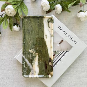 May include: A rectangular tray with a green and white marbled pattern. The tray is resting on a white book titled "The Art of Home." White flowers and green leaves are arranged around the tray.