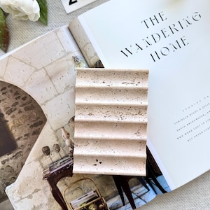 May include: A beige, rectangular soap dish with a ridged design sits on an open book titled "THE WANDERING HOME." The book's pages are cream-colored, and the text is black. A white flower and a tag with the number "212" are also visible.