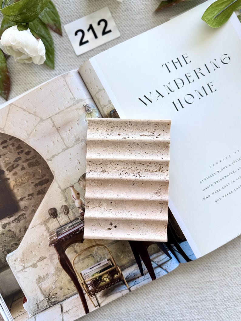 May include: A beige, rectangular soap dish with a ridged design sits on an open book titled "THE WANDERING HOME." The book's pages are cream-colored, and the text is black. A white flower and a tag with the number "212" are also visible.