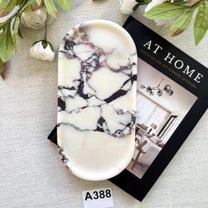 Op de afbeelding: Een ovaalvormig, wit marmeren dienblad met donker bordeauxkleurige adering. De lade heeft afgeronde randen en wordt getoond met een boek getiteld "AT HOME" en decoratieve witte bloemen en groen. Een decoratief woonaccessoire.