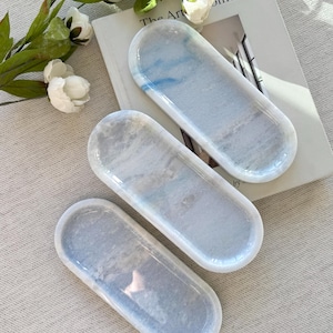 May include: Three oval-shaped trays with a marbled design in shades of white, gray, and blue. The trays are arranged on a neutral-colored surface, with a book and floral accents in the background.