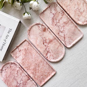 May include: Four rectangular trays with rounded edges, each featuring a pink and white marble pattern. The trays have a smooth, polished surface with distinct veining in shades of deep rose and soft blush, arranged in a diagonal formation.