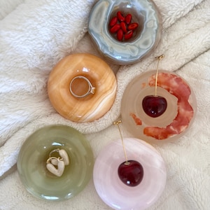 Peut inclure: Cinq présentoirs à bagues décoratifs de différentes couleurs et motifs. L'un contient une bague argentée, un autre des graines rouges, et deux présentent des cerises. Le cinquième contient des boucles d'oreilles en forme de cœur. Les présentoirs sont sur une surface blanche texturée.