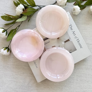 May include: Three round, pale pink glass bowls with a smooth, translucent finish and subtle swirling patterns. The bowls are arranged on top of a white book titled The Art of Home by McGee, accompanied by a decorative branch with white flowers.