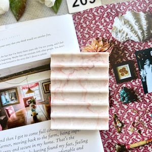 May include: A rectangular, light pink marble soap dish with a ridged design. The dish has subtle pink veining throughout. The number "209" is visible in the background, along with a book and decorative elements.