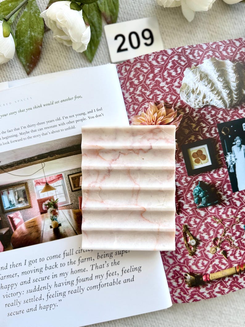 May include: A rectangular, light pink marble soap dish with a ridged design. The dish has subtle pink veining throughout. The number "209" is visible in the background, along with a book and decorative elements.
