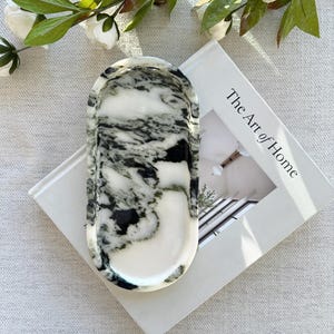 Puede incluir: Una bandeja decorativa ovalada con un diseño de mármol en negro, blanco y gris. La bandeja está sobre un libro titulado "The Art of Home". Hay vegetación con flores blancas en el fondo.