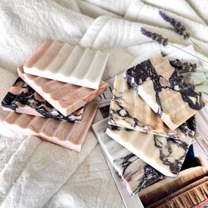 May include: A collection of marble soap dishes in various colors, including pink, white, and black. Each dish has a ridged design for soap drainage. The dishes are arranged on a white, textured surface, with a magazine and lavender sprigs in the background.