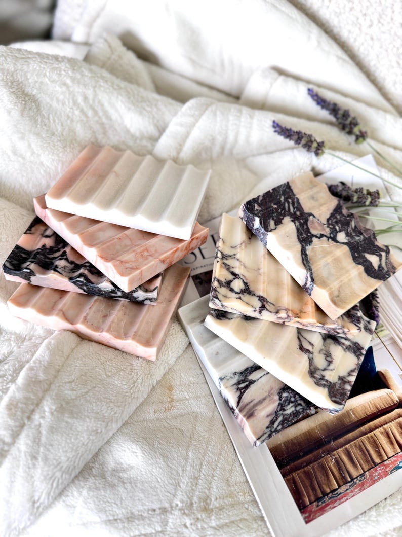 May include: A collection of marble soap dishes in various colors, including pink, white, and black. Each dish has a ridged design for soap drainage. The dishes are arranged on a white, textured surface, with a magazine and lavender sprigs in the background.