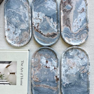 May include: Five oval-shaped trays made of blue and grey stone with a marbled pattern featuring swirls of white and brown. Each tray has a smooth, polished surface and a raised edge. The trays are arranged in a group next to a book titled The Art of Home.