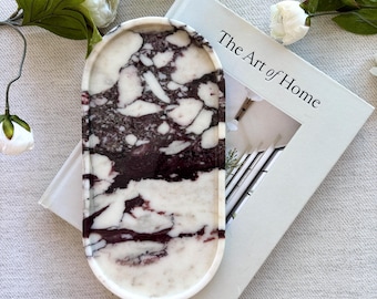 May include: An oval tray featuring a polished surface with a marbled pattern of deep purple and white veins. The tray is positioned on top of a white book titled The Art of Home, surrounded by white artificial flowers with green leaves.