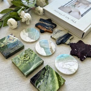 May include: A collection of decorative coasters in various shapes and colors. The coasters are made of stone and feature a range of patterns, including green, white, and blue swirls. Some are square, round, or star-shaped. A book titled "The Art of Home" is in the background.