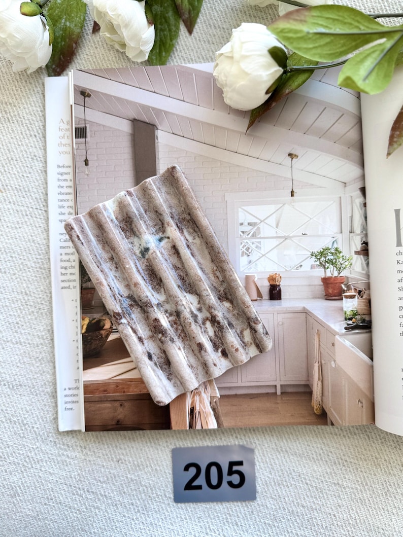 May include: A rectangular, corrugated decorative piece with a weathered, textured appearance. The piece features shades of brown, gray, and white, resembling aged metal. The number "205" is visible on a gray label. The piece is resting on an open magazine.