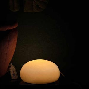May include: A glowing, egg-shaped lamp with a warm, yellow light. The lamp sits on a dark surface, with a tag visible on the left. The background is a deep, dark color.