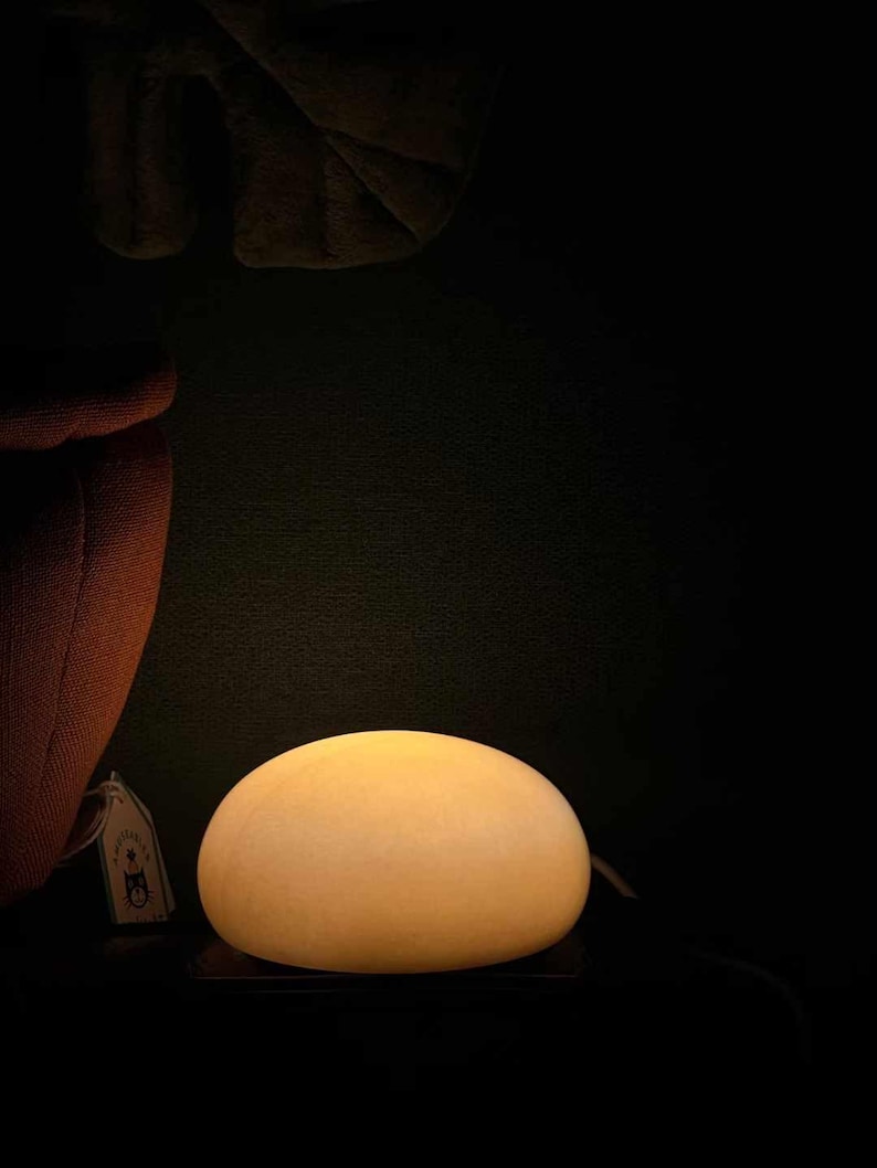 May include: A glowing, egg-shaped lamp with a warm, yellow light. The lamp sits on a dark surface, with a tag visible on the left. The background is a deep, dark color.