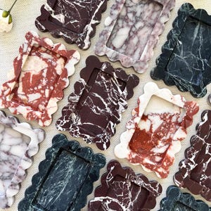 May include: A collection of rectangular trays with scalloped edges, crafted from various types of marble. The trays feature distinct veining patterns in shades of deep burgundy, dark green, white, and reddish-brown, each with a smooth finish.