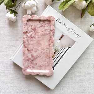 May include: A rectangular pink marble tray with a scalloped edge design. The tray features natural white and dark pink veining throughout its surface. It is positioned on top of a white book titled The Art of Home, surrounded by white flowers.