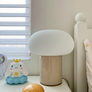 May include: A mushroom-shaped table lamp with a white shade and a beige base sits on a white nightstand. A cartoon character figurine and a small, orange, rounded object are also on the nightstand. The background includes a window with blinds and a white bed frame.